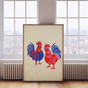 Op de afbeelding: Een ingelijste print van twee hanen, een blauwe en een rode, met witte achtergronden. De hanen staan ​​tegenover elkaar en hebben hun vleugels uitgespreid.