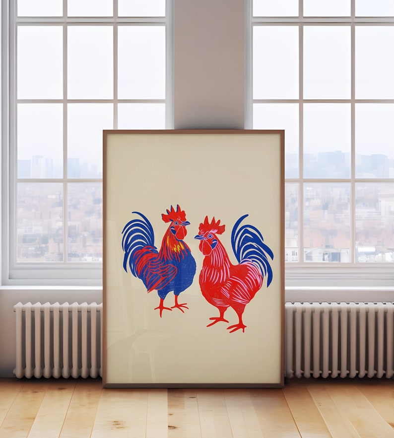 Op de afbeelding: Een ingelijste print van twee hanen, een blauwe en een rode, met witte achtergronden. De hanen staan ​​tegenover elkaar en hebben hun vleugels uitgespreid.