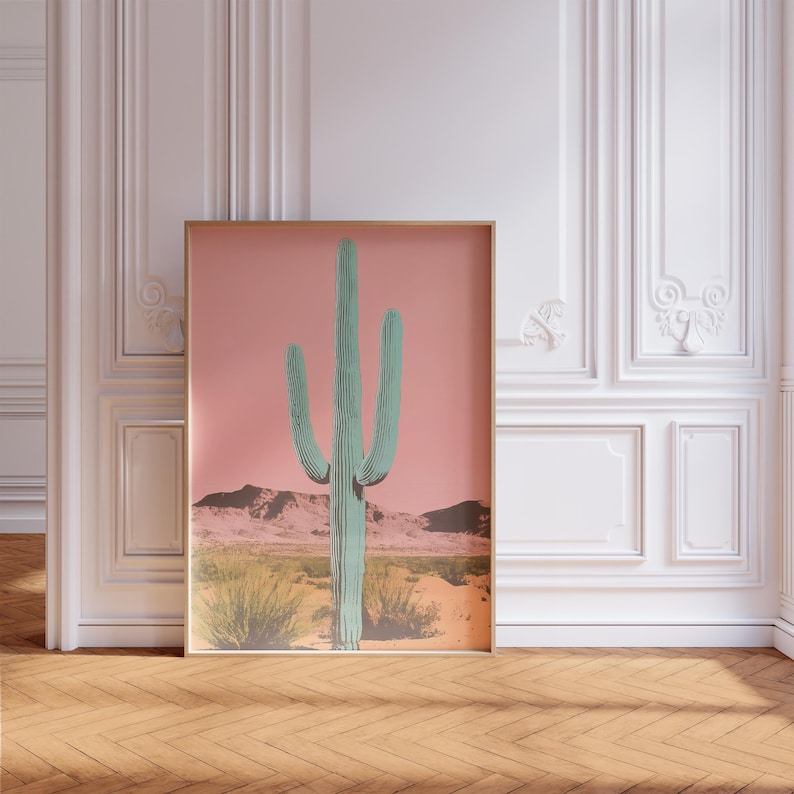 May include: A framed print of a green cactus against a pink background. The cactus is in a desert landscape with hills in the background.