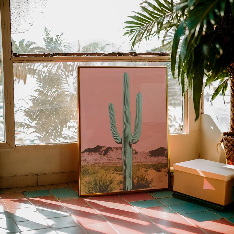 May include: A framed print of a green cactus against a pink background. The cactus is in a desert landscape with sand dunes and small plants.