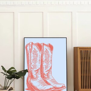 May include: A framed print of a pair of pink cowboy boots on a light blue background. The boots have intricate designs and are set against a white wall.