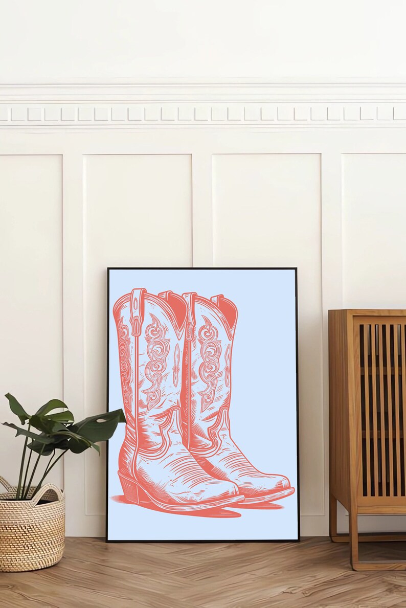 May include: A framed print of a pair of pink cowboy boots on a light blue background. The boots have intricate designs and are set against a white wall.