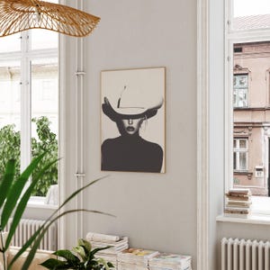May include: Black and white photo print of a woman wearing a large white cowboy hat. The woman is looking directly at the viewer. The photo is framed in a thin, light wood frame.