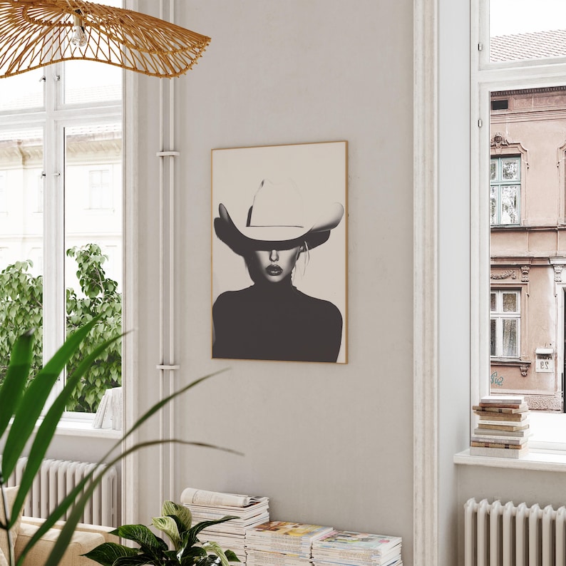 May include: Black and white photo print of a woman wearing a large white cowboy hat. The woman is looking directly at the viewer. The photo is framed in a thin, light wood frame.