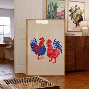 Op de afbeelding: Een ingelijste print van twee rode en blauwe hanen met een witte achtergrond. De hanen staan ​​tegenover elkaar en hebben hun vleugels uitgespreid. De print hangt aan een muur in een kamer met een houten vloer.