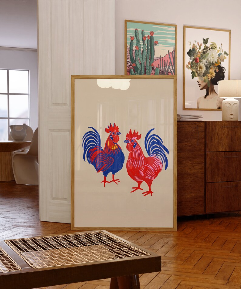 Op de afbeelding: Een ingelijste print van twee rode en blauwe hanen met een witte achtergrond. De hanen staan ​​tegenover elkaar en hebben hun vleugels uitgespreid. De print hangt aan een muur in een kamer met een houten vloer.