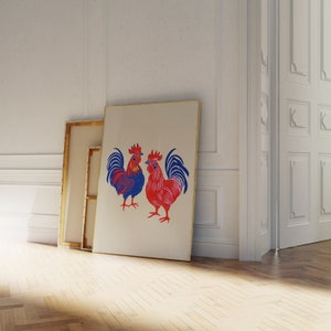 Op de afbeelding: Een ingelijste print van twee rode en blauwe hanen met zwarte contouren. De hanen staan ​​tegenover elkaar en hebben hun vleugels uitgespreid. De print is op een witte achtergrond en is ingelijst in een gouden lijst.