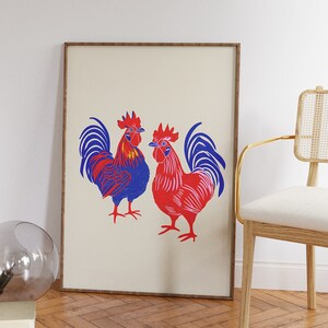 Op de afbeelding: Een ingelijste print van twee hanen, een blauwe en een rode, met witte achtergronden. De hanen staan ​​tegenover elkaar en hebben hun vleugels uitgespreid.