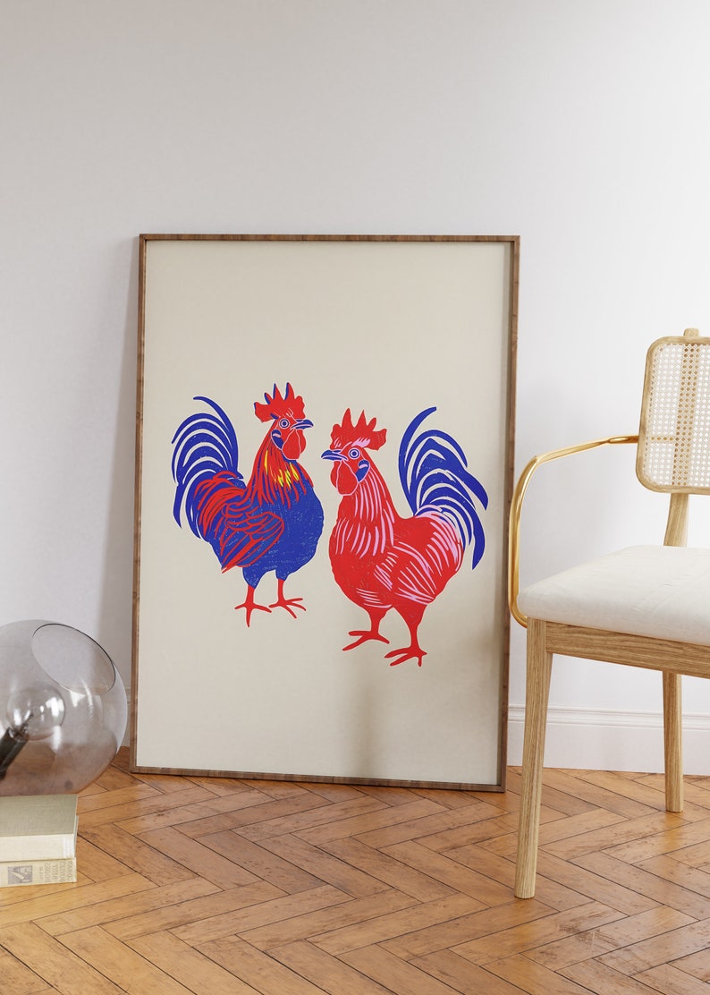 Op de afbeelding: Een ingelijste print van twee hanen, een blauwe en een rode, met witte achtergronden. De hanen staan ​​tegenover elkaar en hebben hun vleugels uitgespreid.