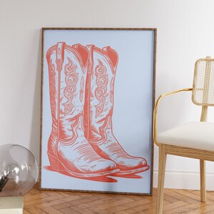 May include: A framed print of a pair of coral-colored cowboy boots on a light blue background.