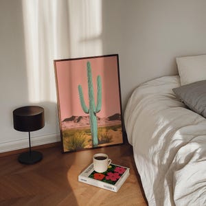 May include: A framed print of a green cactus against a pink background. The print is sitting on a table next to a book and a cup of coffee.