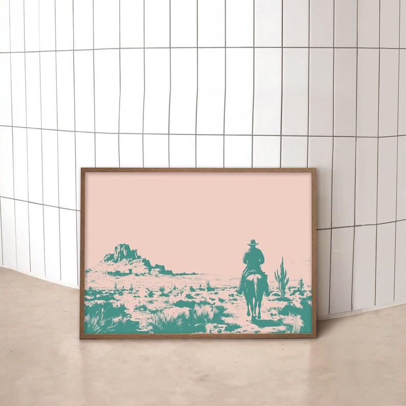 Op de afbeelding: Een ingelijst drukwerk van een cowboy die op een paard door een woestijnlandschap rijdt. De print is in een houten lijst en heeft een roze achtergrond met turquoise details.