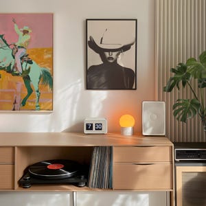 May include: A wooden cabinet with two drawers and a shelf. The cabinet has a record player, a stack of vinyl records, a small clock that reads "7 30", a lamp with an orange globe, and a white speaker on top. There are two framed pictures on the wall behind the cabinet.