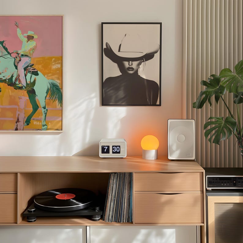 May include: A wooden cabinet with two drawers and a shelf. The cabinet has a record player, a stack of vinyl records, a small clock that reads "7 30", a lamp with an orange globe, and a white speaker on top. There are two framed pictures on the wall behind the cabinet.
