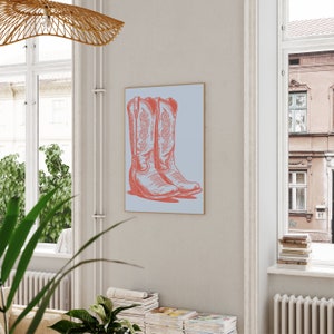 May include: A framed print of a pair of pink cowboy boots with intricate designs on the front. The boots are set against a light blue background.