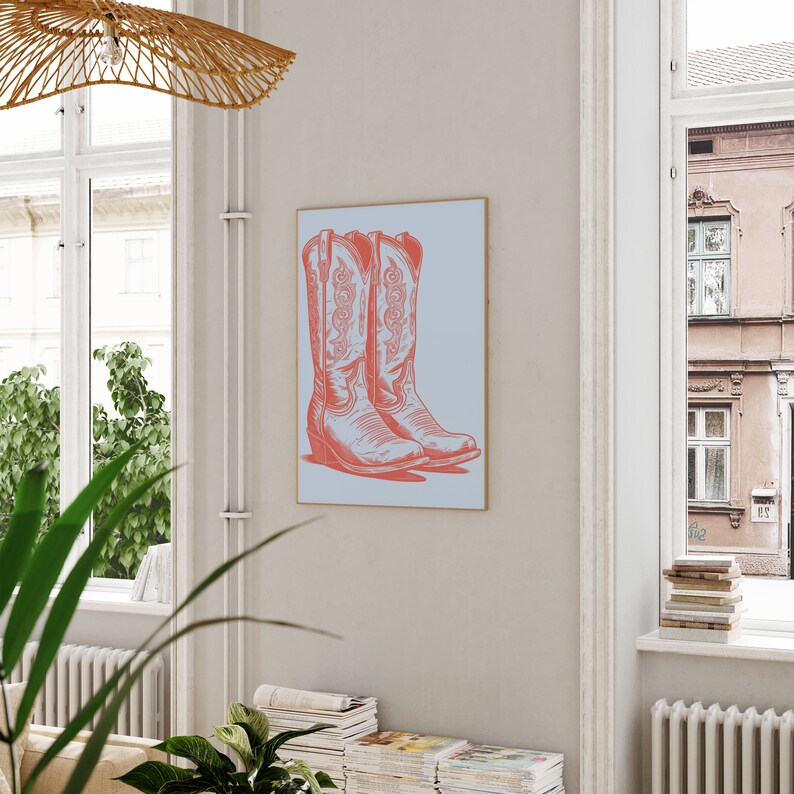 May include: A framed print of a pair of pink cowboy boots with intricate designs on the front. The boots are set against a light blue background.