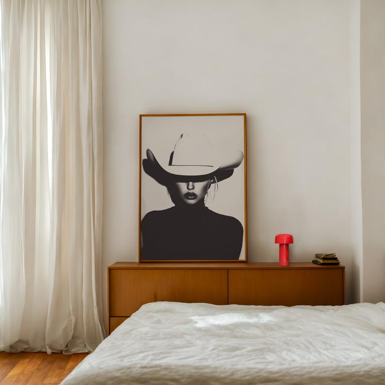 May include: A black and white photograph of a woman wearing a white cowboy hat. The photograph is framed and hanging on a white wall above a wooden dresser with a red lamp and a book on top. A white bed with a white linen duvet is in the foreground.