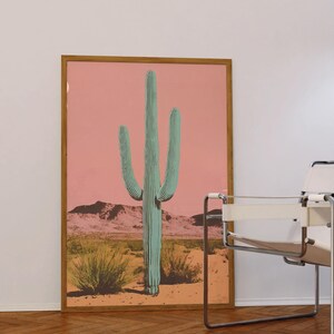 May include: A framed print of a large green cactus in a desert landscape with a pink sky. The cactus is the focal point of the image and is surrounded by smaller plants and a mountain range in the background.