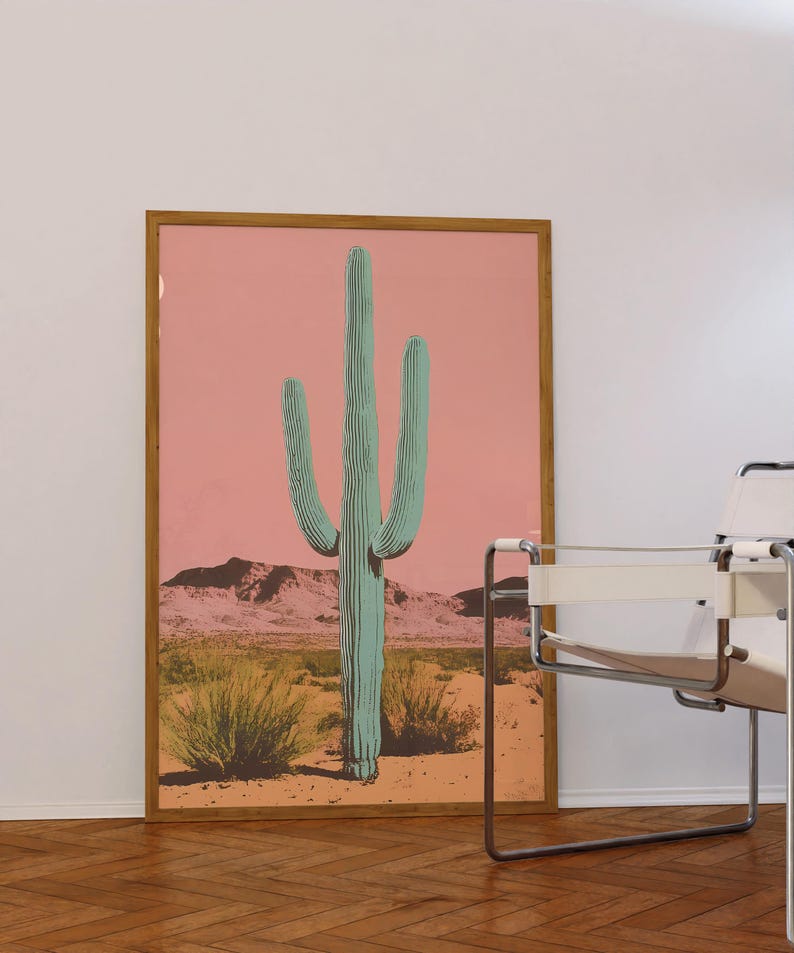 May include: A framed print of a large green cactus in a desert landscape with a pink sky. The cactus is the focal point of the image and is surrounded by smaller plants and a mountain range in the background.