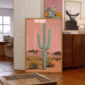 May include: A framed print of a tall, green cactus in a desert landscape with a pink sky. The cactus is in the center of the image and is surrounded by smaller plants and mountains in the background.