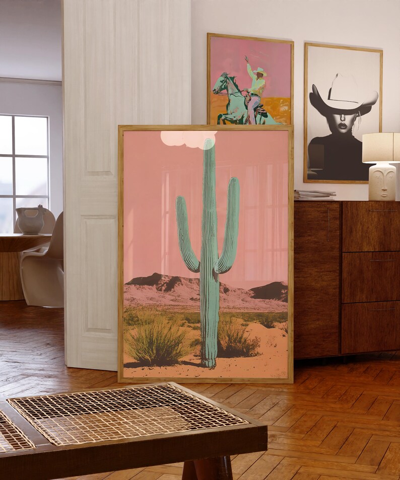 May include: A framed print of a tall, green cactus in a desert landscape with a pink sky. The cactus is in the center of the image and is surrounded by smaller plants and mountains in the background.