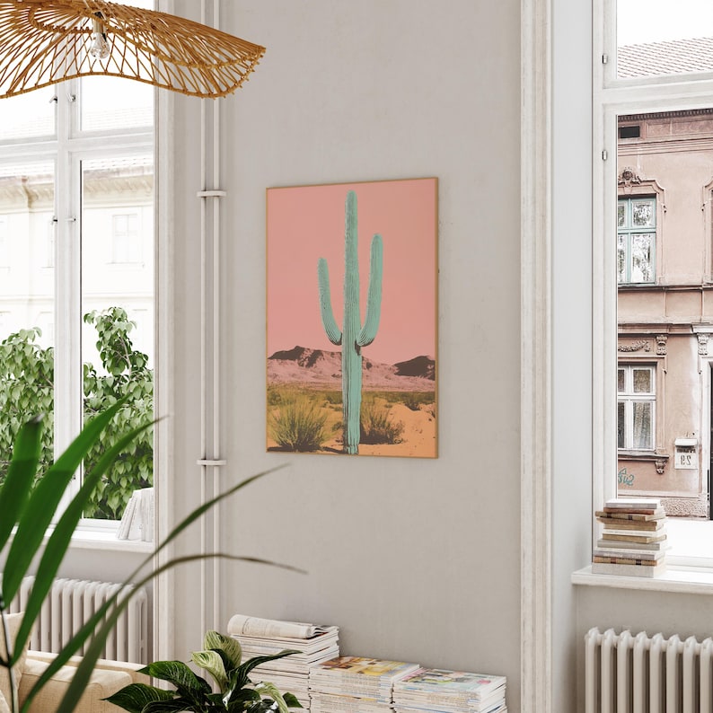 May include: A digital art print featuring a large, teal-colored cactus in a desert landscape with a pink sky. The print is framed and hanging on a white wall.