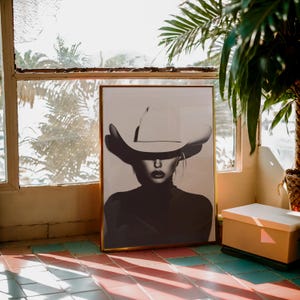 May include: A black and white photograph of a person wearing a large white cowboy hat. The person is looking directly at the camera. The photograph is framed in a gold frame.