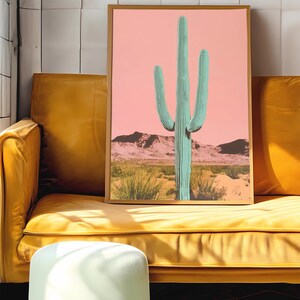 May include: A framed print of a green cactus in a desert landscape with a pink sky. The print is in a gold frame and is hanging on a wall.