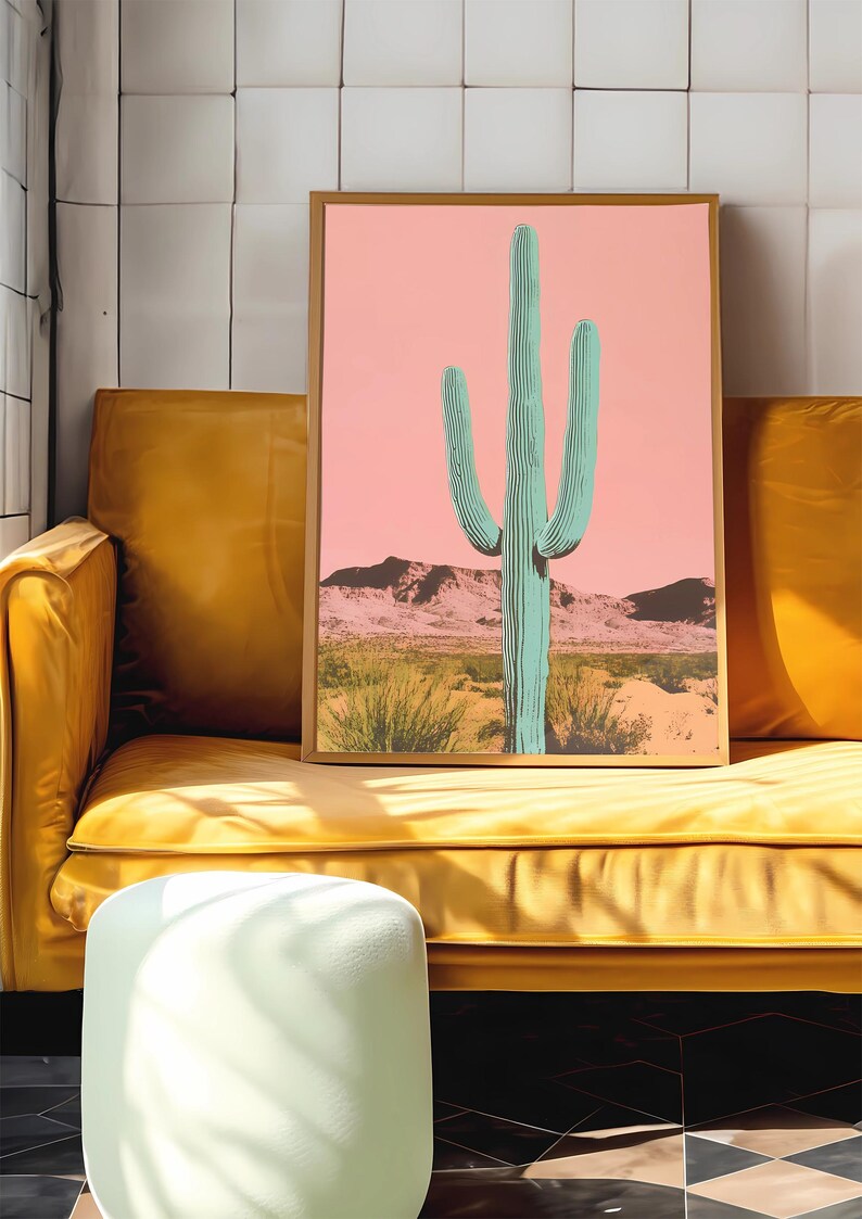 May include: A framed print of a green cactus in a desert landscape with a pink sky. The print is in a gold frame and is hanging on a wall.