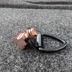 May include: A black metal D-ring tie-down anchor featuring a silver-colored hexagonal bolt and a circular washer. The hardware component has a triangular loop attached to a square-shaped base, with red outlines highlighting the bolt and base structure.
