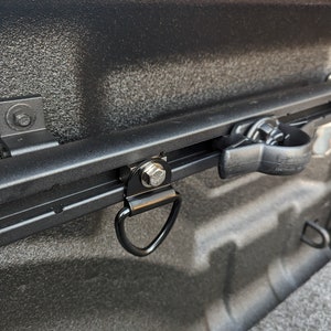 May include: A black metal D-ring with a silver screw is attached to a black metal track. The track is mounted to the inside of a truck bed.