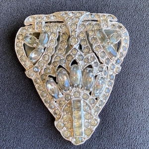 Può includere: Spilla vintage color argento con un design simmetrico. La spilla è incastonata con numerosi strass chiari e scintillanti. La forma è un design traforato stilizzato con una forma centrale allungata.