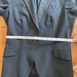 Op de afbeelding: Een donkergrijze blazer met een enkele knoopsluiting, twee voorzakken en een revers. De blazer wordt gemeten met een meetlint, met een taillemeting van ongeveer 38 cm.