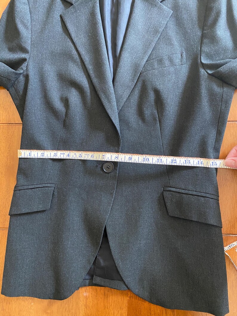 Op de afbeelding: Een donkergrijze blazer met een enkele knoopsluiting, twee voorzakken en een revers. De blazer wordt gemeten met een meetlint, met een taillemeting van ongeveer 38 cm.