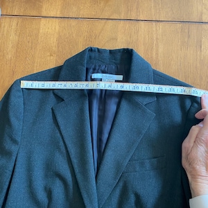 Op de afbeelding: Een donkergrijze blazer met een revers, een sluiting met twee knopen en een borstzak. De blazer wordt gemeten met een liniaal. Het label luidt "ZARA BASIC".