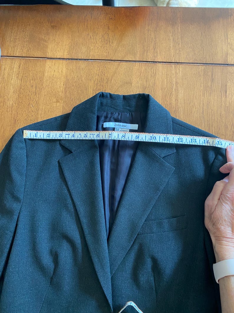 Op de afbeelding: Een donkergrijze blazer met een revers, een sluiting met twee knopen en een borstzak. De blazer wordt gemeten met een liniaal. Het label luidt "ZARA BASIC".