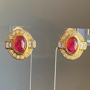 以下が含まれることがあります： ピンクの楕円形のcabochonストーンがクリアなラインストーンのハローで囲まれた、ゴールドトーンのイヤリングのペア。イヤリングは、楕円形のストーンの両側に正方形のラインストーンが付いています。