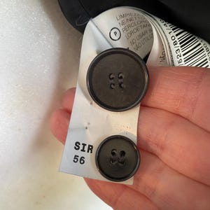 Op de afbeelding: Close-up van twee donkergrijze knopen bevestigd aan een wit label. Het label heeft de tekst "SIR 56" en tekst in meerdere talen, waaronder "LIMPIEZA" en "N&Atilde;O UTILIZ".