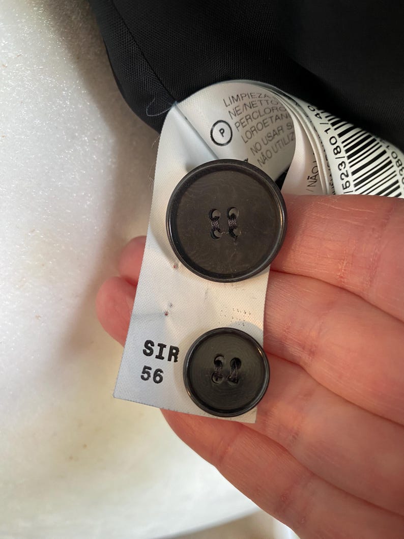 Op de afbeelding: Close-up van twee donkergrijze knopen bevestigd aan een wit label. Het label heeft de tekst "SIR 56" en tekst in meerdere talen, waaronder "LIMPIEZA" en "N&Atilde;O UTILIZ".