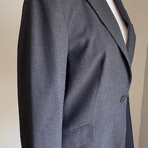 Op de afbeelding: Een antracietgrijze blazer gepresenteerd op een witte mannequin. De blazer heeft een revers, een sluiting met &eacute;&eacute;n knoop en een voorzak. De mouwen hebben knopen bij de manchetten. De stof lijkt een geweven materiaal te zijn.