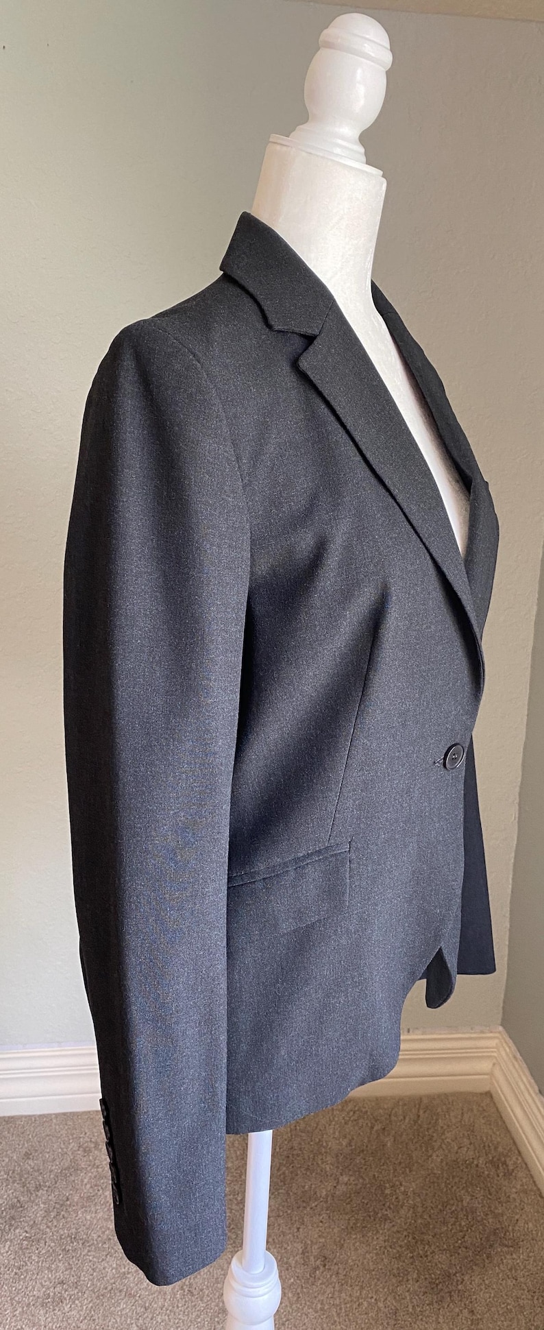 Op de afbeelding: Een antracietgrijze blazer gepresenteerd op een witte mannequin. De blazer heeft een revers, een sluiting met &eacute;&eacute;n knoop en een voorzak. De mouwen hebben knopen bij de manchetten. De stof lijkt een geweven materiaal te zijn.