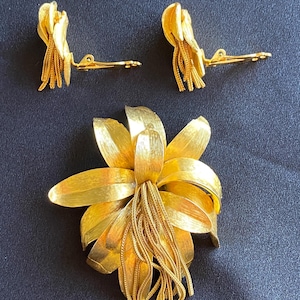 Könnte beinhalten: Ein goldfarbenes Schmuckset mit einer großen Blumenbrosche und zwei passenden Clip-Ohrringen. Die Brosche hat geschichtete Blütenblätter und eine Quaste aus filigranen Ketten. Die Ohrringe haben ebenfalls ein Blumendesign mit Kettenquasten. Das Set wird auf dunklem Hintergrund präsentiert.