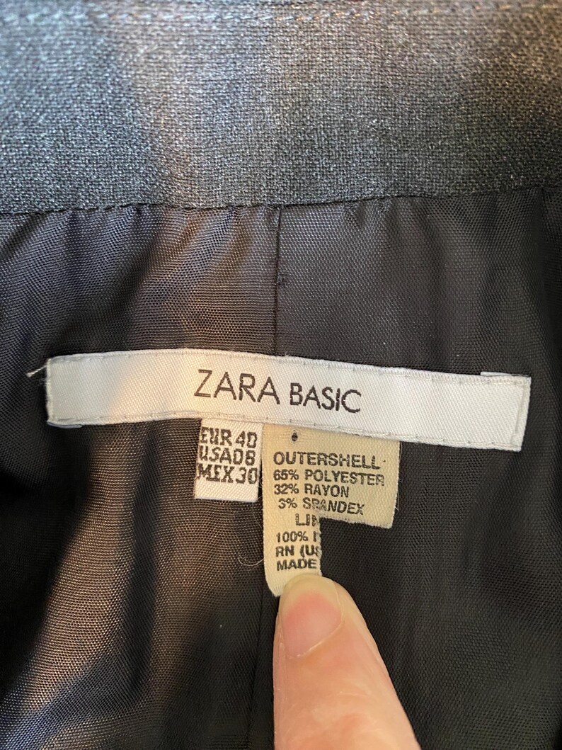 Op de afbeelding: Close-up van de voering van een donkergrijze Zara Basic blazer. Het label toont maat EUR 40, US AD8, MEX 30, en de samenstelling van de buitenkant: 65% polyester, 32% rayon en 3% spandex. De voering is 100% linnen.