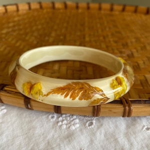 Puede incluir: Brazalete de color crema con flores amarillas secas y hojas marrones incrustadas. El brazalete está colocado sobre una bandeja de bambú tejido y tela de encaje blanco.