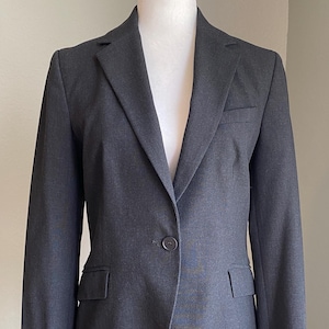 Op de afbeelding: Een donkergrijze blazer op een witte mannequin. De blazer heeft een revers, een sluiting met &eacute;&eacute;n knoop en twee voorzakken. De stof is effen van kleur, geschikt voor zakelijke of formele gelegenheden. De blazer is ongeveer 76 cm lang.
