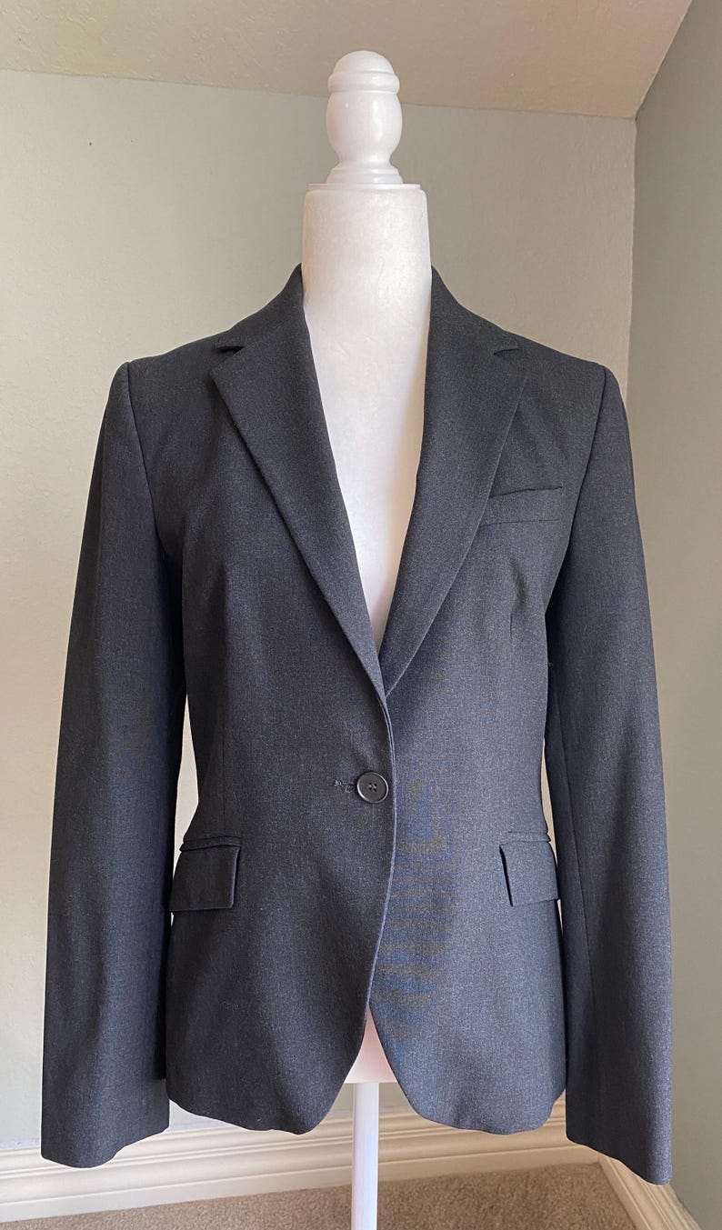 Op de afbeelding: Een donkergrijze blazer op een witte mannequin. De blazer heeft een revers, een sluiting met &eacute;&eacute;n knoop en twee voorzakken. De stof is effen van kleur, geschikt voor zakelijke of formele gelegenheden. De blazer is ongeveer 76 cm lang.