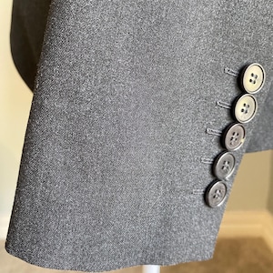 Op de afbeelding: Close-up van een donkergrijze blazer mouw. De stof heeft een subtiele textuur. Vier ronde, grijze knopen zijn zichtbaar op de manchet. De knopen hebben een ontwerp met twee gaten. De blazer is waarschijnlijk gemaakt van een geweven materiaal.