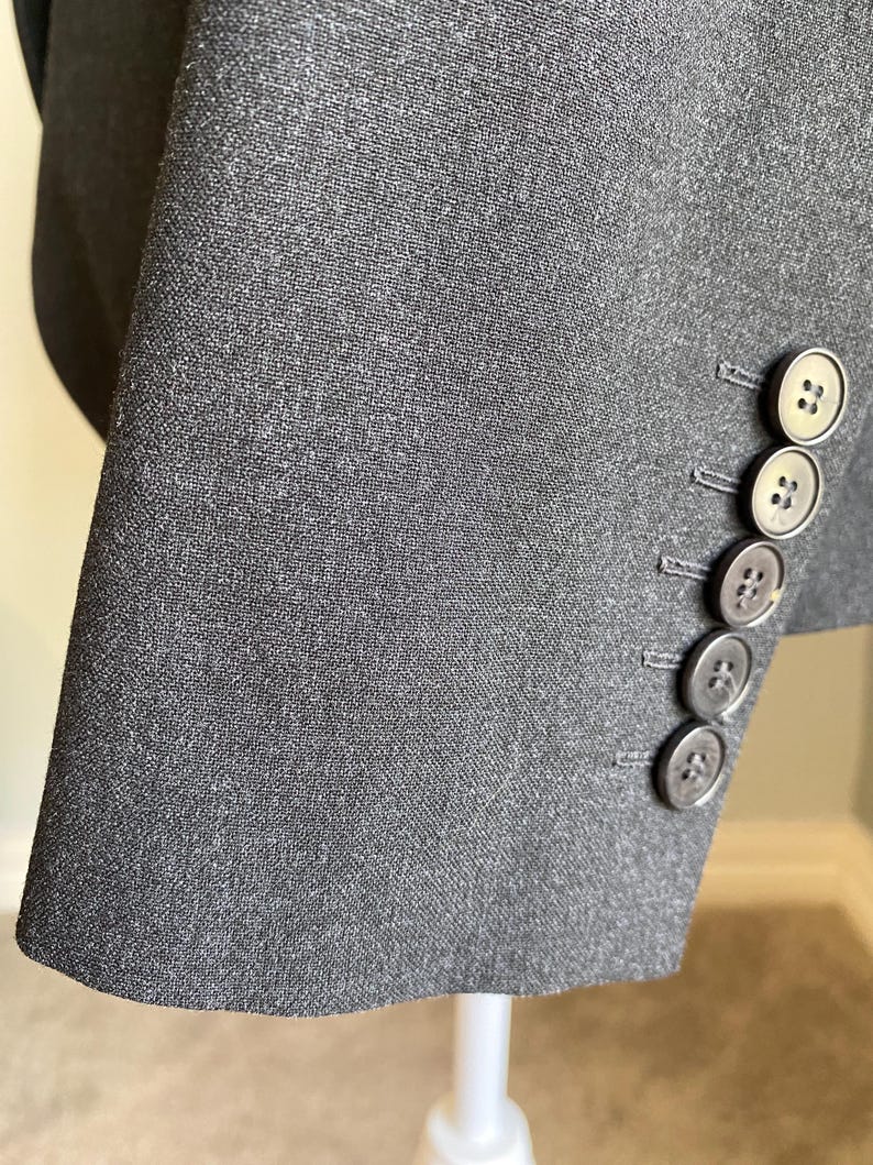 Op de afbeelding: Close-up van een donkergrijze blazer mouw. De stof heeft een subtiele textuur. Vier ronde, grijze knopen zijn zichtbaar op de manchet. De knopen hebben een ontwerp met twee gaten. De blazer is waarschijnlijk gemaakt van een geweven materiaal.