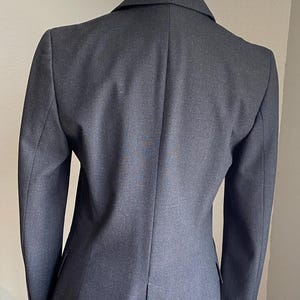 Op de afbeelding: Een donkergrijze blazer op een mannequin. De getailleerde jas heeft een gekerfd revers, lange mouwen en een ontwerp met &eacute;&eacute;n rij knopen. De achterkant van de blazer heeft een split aan de onderkant. De stof lijkt een geweven materiaal te zijn.