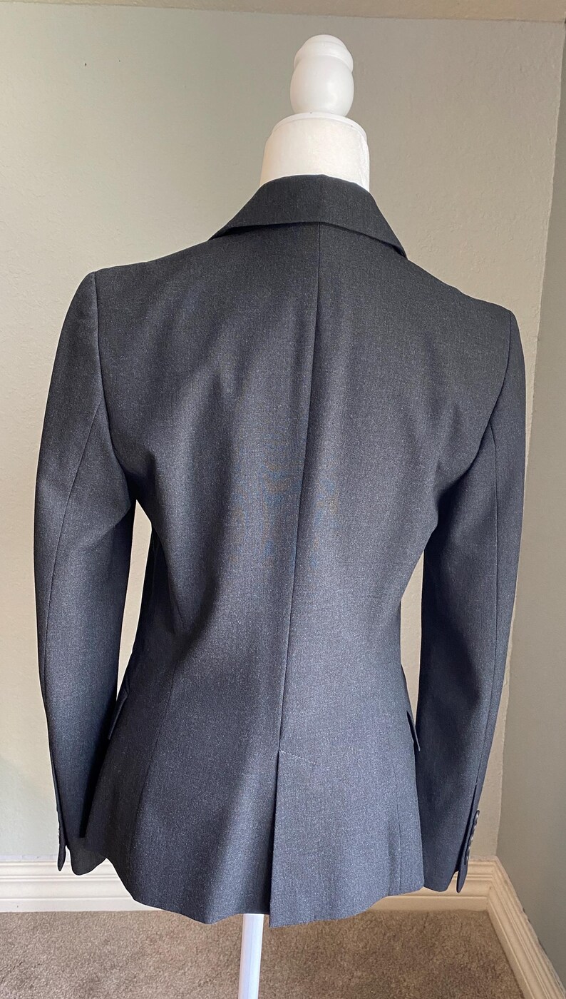Op de afbeelding: Een donkergrijze blazer op een mannequin. De getailleerde jas heeft een gekerfd revers, lange mouwen en een ontwerp met &eacute;&eacute;n rij knopen. De achterkant van de blazer heeft een split aan de onderkant. De stof lijkt een geweven materiaal te zijn.
